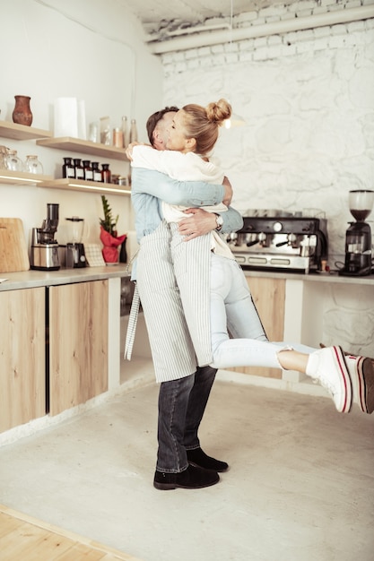 Ámense unos a otros. Mujer feliz saltando en brazos de su guapo marido en su cocina.