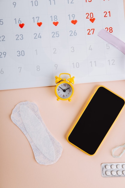 Menstruationskalender und Smartphone mit Damenbinde und gelbem Wecker auf rosa Hintergrund. Frauenkritische Tage, Frauenhygieneschutz.