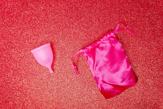 Menstruationstasse mit Satinbeutel auf glänzend rotem Hintergrund Top-View Null-Abfall-Konzept umweltfreundliche Menstruationsprodukte