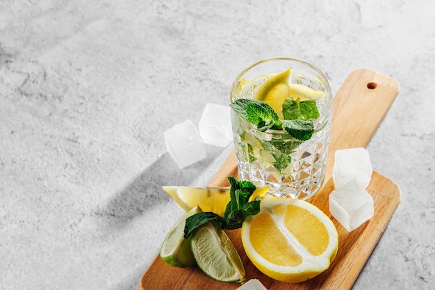 Menta, lima, limones, hielo y utensilios de bar en la tabla de cortar de madera en la vista superior de fondo de mármol blanco