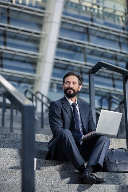 Mentalidad profesional. Agradable empresario barbudo usando su computadora portátil en las escaleras