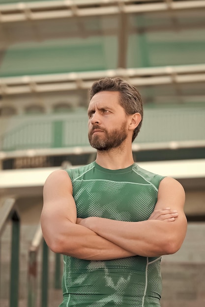 Mente y cuerpo en armonía siente la energía de tus músculos dentro de él estar en forma y flexible entrenador de fitness confiado manos cruzadas hombre serio con barba entrenando al aire libre pequeño descanso en el entrenamiento