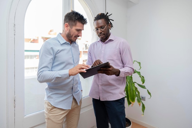 Mentor des afroamerikanischen Managers berät seinen Kollegen über Investitionsoperationen mit einem Tablet-Gerät, das im Büro in der Nähe des Fensters steht