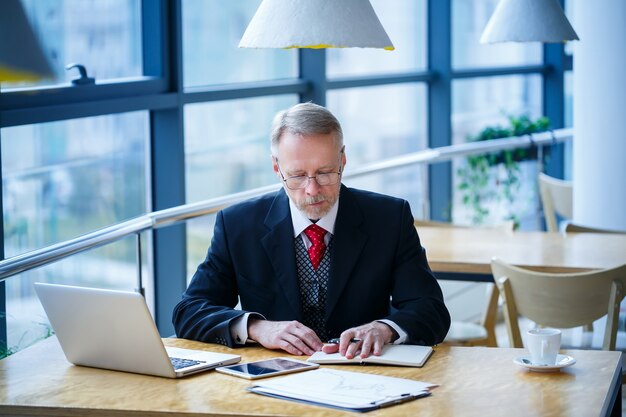 Mentor de sexo masculino adulto, director, empresario con gafas y traje estudiando documentos mientras está sentado en la mesa. Concepto de jornada laboral
