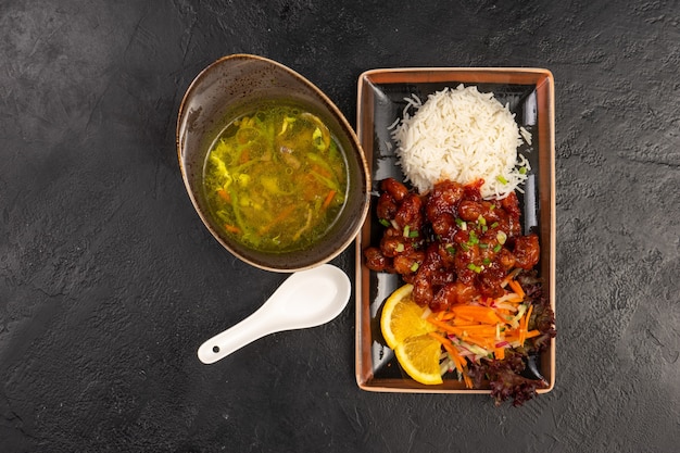 Menú de almuerzo de sopa de verduras y plato de carne caliente con arroz y verduras frescas como guarnición. Dos platos asiáticos en platos tradicionales inusuales sobre una mesa de piedra negra.