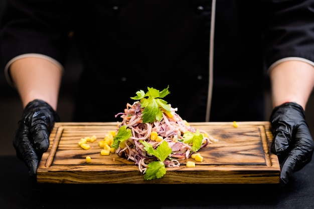 Menu vegetariano do restaurante. Foto recortada do chef em luvas de cozinha pretas, segurando uma placa de madeira com salada de repolho salada de repolho.