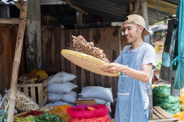 Mercearia asiática segurando uma bandeja de bambu, peneirando açafrão para limpar em uma barraca de legumes