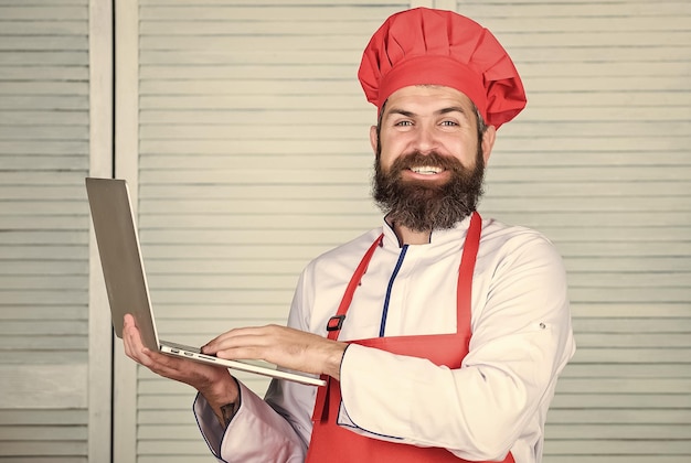 Mercearia on-line Serviço de entrega Laptop de chef na cozinha Escola de culinária Hipster de chapéu e avental comprar produtos on-line Compras on-line Chef masculino pesquisando ingredientes on-line cozinhando comida