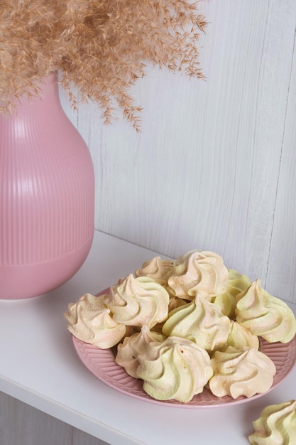 Merengue aireado amarillo sobre un plato rosa cerca de un jarrón con cañas sobre un fondo blanco