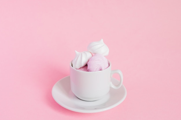 Merengues retorcidos blancos y rosados en una pequeña taza de café de porcelana sobre fondo rosa. Postre francés preparado con batido de azúcar y claras de huevo al horno. Tarjeta de felicitación con espacio de copia