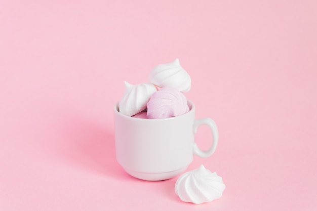 Merengues torcidos brancos e rosa em uma xícara de café de porcelana pequena em fundo rosa. Sobremesa francesa preparada a partir de chantilly com açúcar e claras de ovos cozidas. Cartão com espaço de cópia