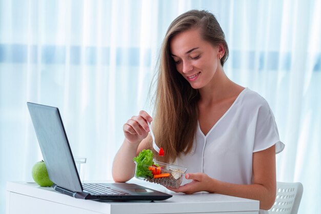 Foto merienda saludable en el lugar de trabajo de oficina. mujer de negocios feliz comiendo comidas de la caja de almuerzo en la mesa de trabajo durante la hora del almuerzo