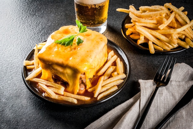 Merienda tradicional portuguesa. Francesinha sandwich de pan, queso, carne de cerdo, jamón, salchichas, con salsa de cerveza de tomate y papas fritas. Con un vaso de cerveza y papas. En mesa negra. Copia espacio