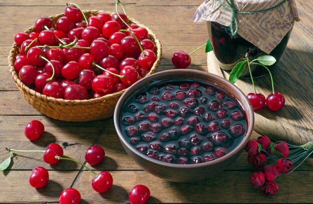 Mermelada de cerezas en un cuenco de arcilla y cerezas rojas maduras en una canasta sobre una mesa de madera. Estilo rústico.