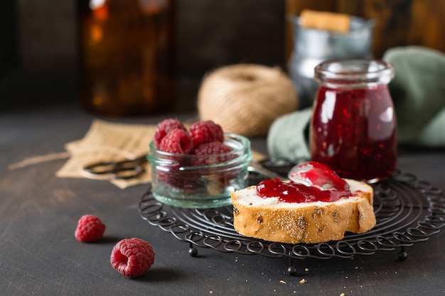 Mermelada de frambuesa en tarro de cristal y sándwich con bayas de frambuesa frescas en el antiguo fondo retro