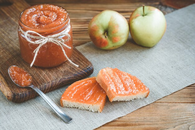 Mermelada de manzana en el banco con manzanas, rebanada de pan con mermelada y cuchara de mermelada en tela de lino sobre fondo de madera.