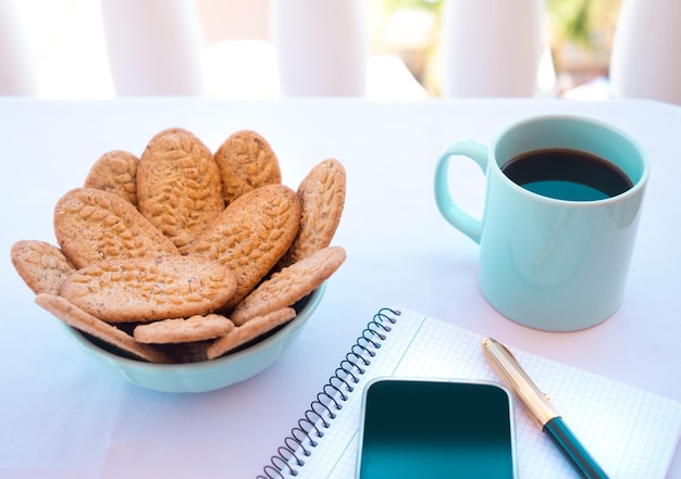 Mesa blanca lista para desayunar o descansar al aire libre en el balcón con taza de café y galletas integrales Teléfono celular e instrumentos de escritura en la mesa