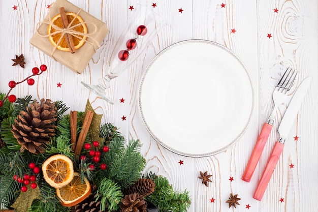 Mesa blanca de Navidad rústica con decoración navideña blanca placa vacía colocación cubiertos copa de vino ramas de abeto conos naranjas secas regalo envuelto en papel kraft
