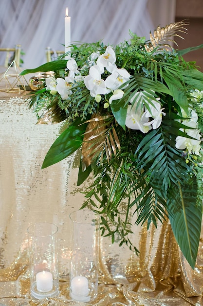 Mesa de bodas con adornos dorados hojas verdes tropicales y velas encendidas en jarrones de vidrio