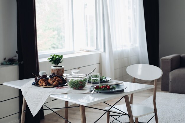 Mesa com saboroso café da manhã saudável, rodeado por um interior moderno na manhã de verão. comida apetitosa fresca, tomate orgânico, hortaliças, croissant e frutas em pratos à mesa nomeações