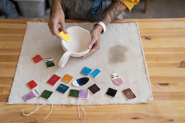 Mesa com tintas para colorir copo de oleiro e mão de designer escolhendo a cor para pintar caneca de cerâmica