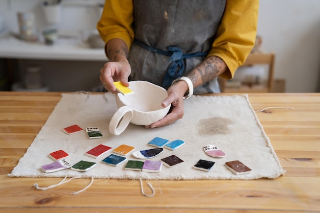 Mesa com tintas para colorir copo de oleiro e mão de designer escolhendo a cor para pintar caneca de cerâmica