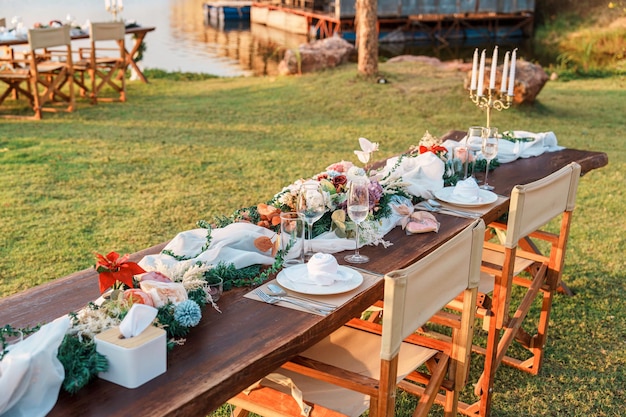 Mesa de comedor de estilo rústico con decoración de velas de copa de vino de flores en la cena junto al río al atardecer