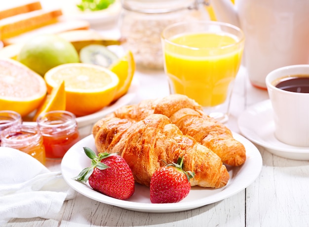 Mesa de café da manhã com croissants, café, suco de laranja, torradas e frutas