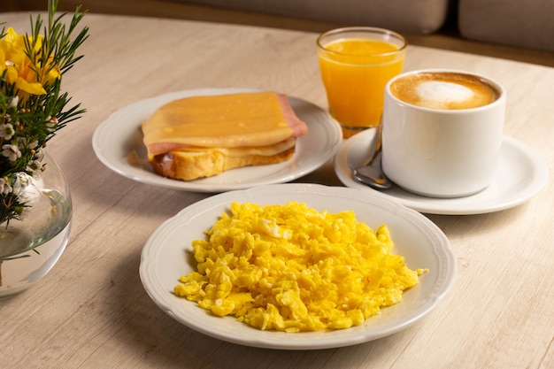 Mesa de café da manhã com ovos, xícara de café, suco de laranja e fatia de pão com queijo e bacon.