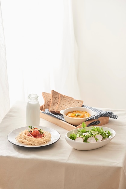 Mesa de café da manhã; espaguete servido com rabanete e salada com purê de abóbora, pão e leite na mesa