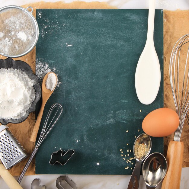 Mesa de cozinha com placa verde em branco, ingredientes de bolo de cozimento (ovos, farinha, leite, cortador de biscoitos, semente de gergelim, açúcar) e utensílios de cozimento ao redor. placa em branco de fundo com espaço de texto de receita grátis.