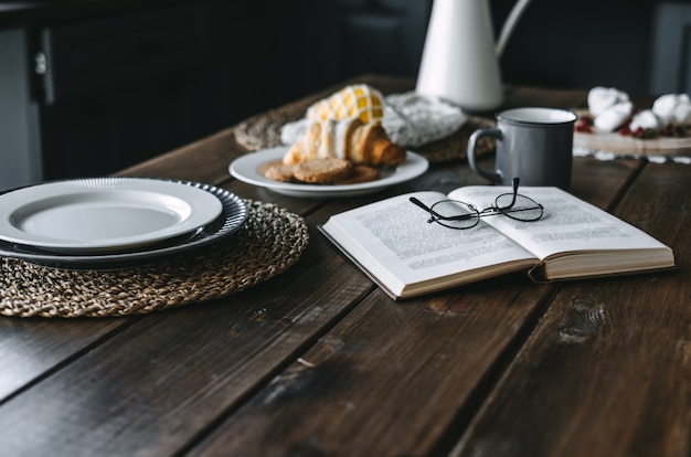 Mesa de cozinha de madeira com café da manhã, livro, xícara de café e croissant.