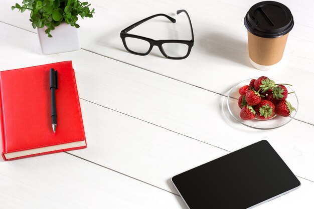 Mesa de escritório com conjunto de suprimentos bloco de notas vermelho copo caneta tablet óculos flor em fundo branco ...