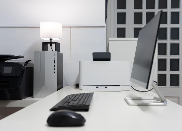 Mesa de escritório com um pc preto e cinza, impressora preto e branco e uma tela