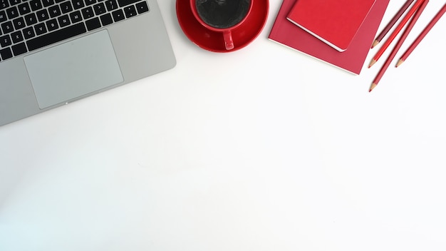 Mesa de escritório de vista superior com computador portátil, xícara de café vermelha, caderno vermelho e lápis na mesa branca.