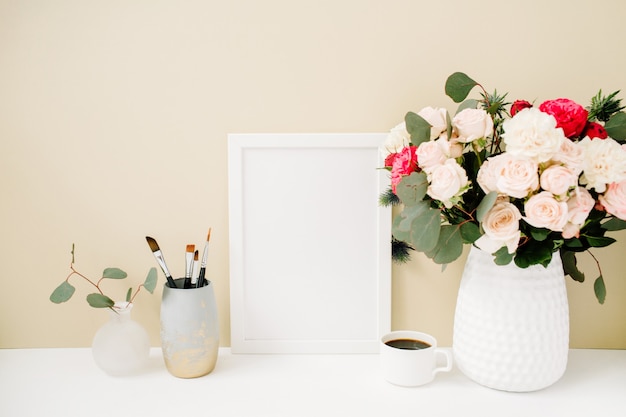 Mesa de escritório em casa com simulação de moldura de foto, lindas rosas e buquê de eucalipto na frente de um fundo bege claro