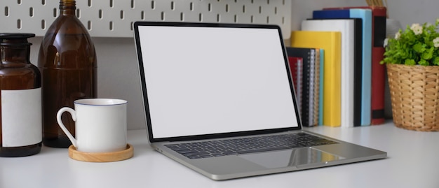 Mesa de escritório em casa elegante com laptop de tela em branco, livros, xícara e decorações na mesa branca