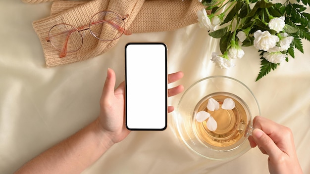 Mesa de escritório feminina com a mão de uma mulher segurando uma maquete de smartphone e uma xícara de chá. tela em branco do telefone.