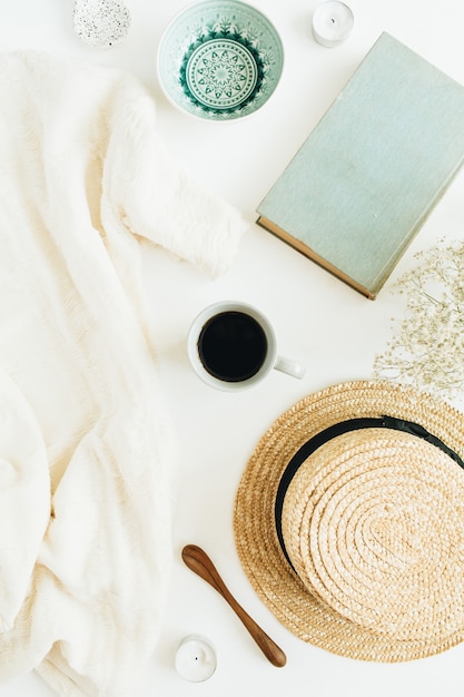 Mesa de escritório feminina com café, livro, chapéu de palha, flores e cobertor na superfície branca