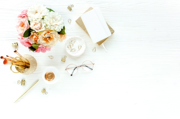 Mesa de escritório feminina plana Espaço de trabalho feminino com rosas cor-de-rosa flores bouquet acessórios óculos de diário dourados em fundo branco de madeira Vista superior