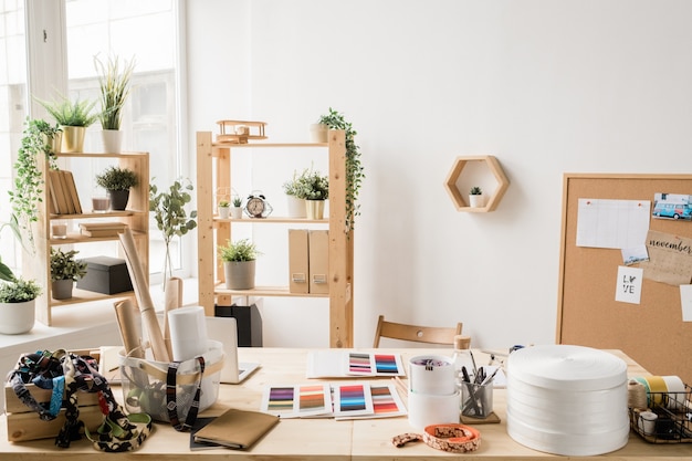 Mesa de madeira à janela com vários materiais para o trabalho criativo de designer de moda ou de interiores em um estúdio ou escritório moderno