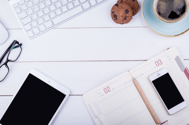 Mesa de madeira branca com tablet, caderno, café preto, caneta, telefone, teclado, biscoito e copos. Ângulo de visão superior com espaço de cópia