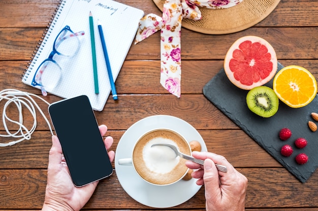 Mesa de madeira posta para um café da manhã saudável. Cappuccino quente e frutas cítricas. Mão humana segura um celular