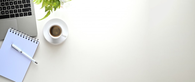 Mesa de trabalho branca que cercada por computador portátil, caneta, nota, planta em vaso e xícara de café.