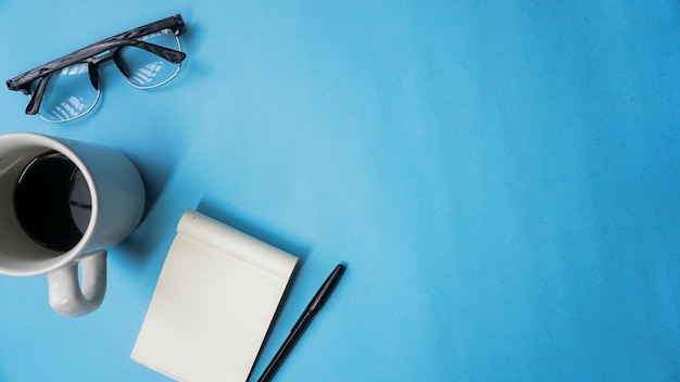 mesa de trabalho com cadernos, canetas, óculos e café em fundo azul