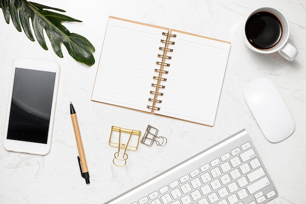 Mesa de trabalho elegante com páginas de caderno em branco, teclado, óculos, smartphone e caneca de café na mesa de mármore