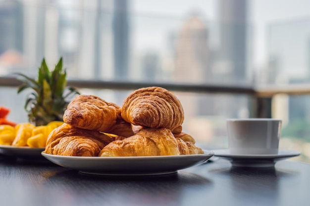 Mesa de desayuno con café, fruta y croissant de pan en un balcón con el telón de fondo de la gran ciudad