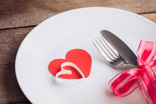 Mesa de día de San Valentín con plato, cuchillo, tenedor, cinta roja y corazones en mesa de madera