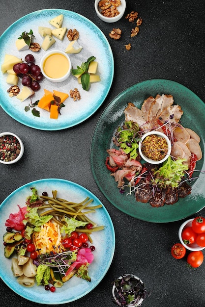 Mesa de embutidos con tres platos en la vista superior de fondo de hormigón oscuro. plato de queso con miel y uva, antipasti de verduras en escabeche, plato de jamón