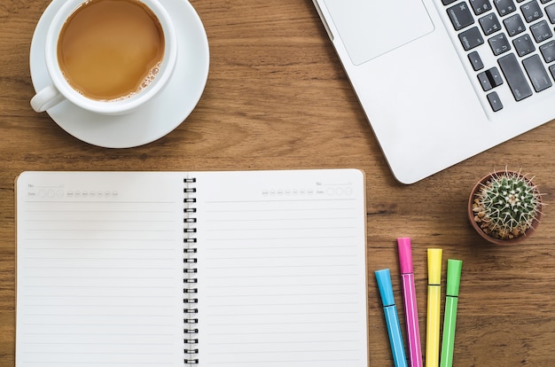 Mesa de escritorio de madera con el cuaderno, la computadora portátil, la pluma del color, el cactus y la taza de café. Vista superior con espacio de copia.